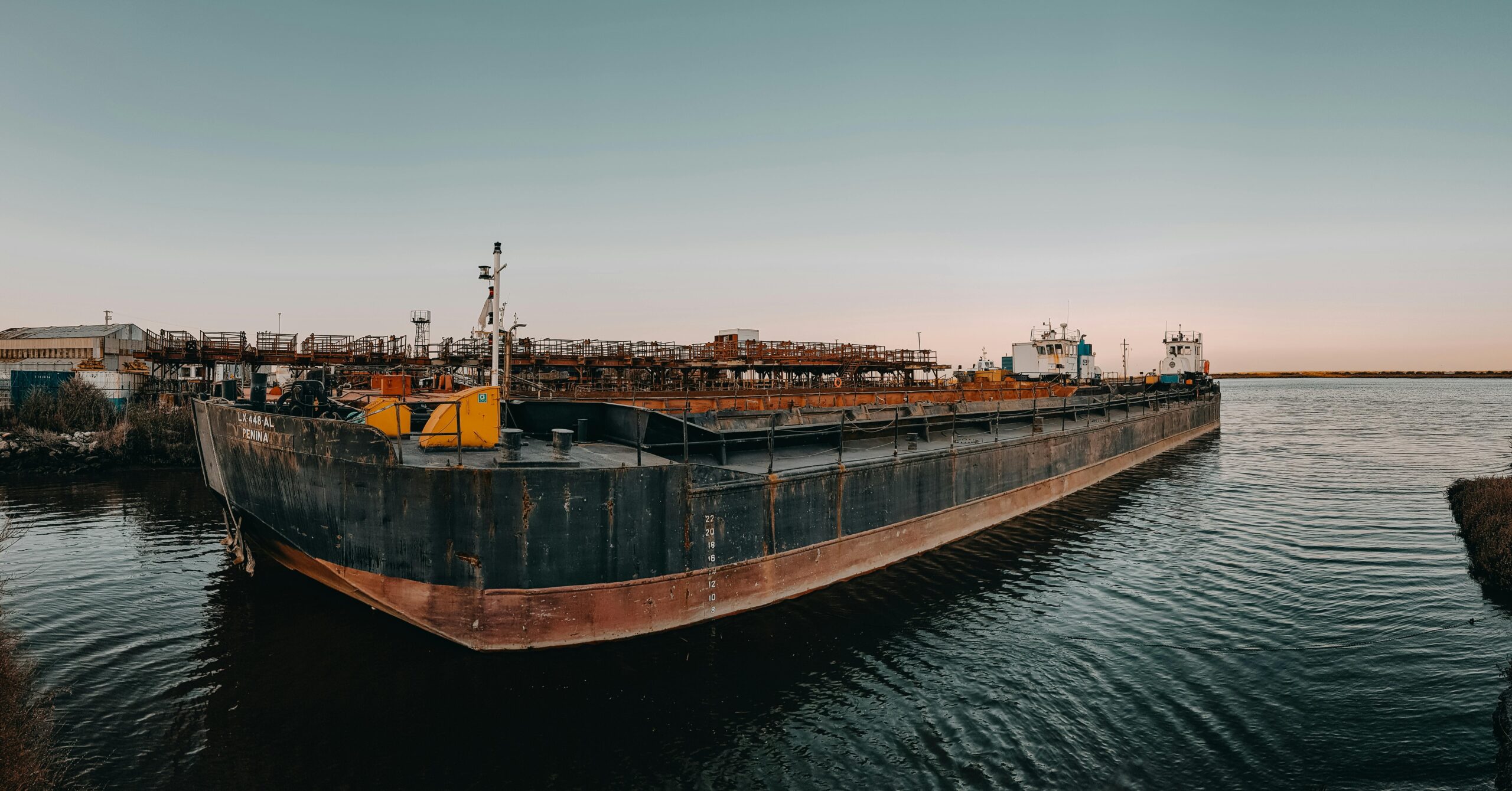 A large cargo barge docked at an industrial harbor during sunset, calm waters and clear sky.